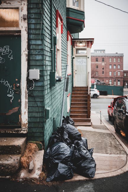 A group of black plastic rubbish bags stacked against the green-painted exterior wall of a building on a pavement, near the bottom of a wooden staircase leading to a white door with a small awning. The building has teal-colored horizontal weatherboard cladding with red and white trim around windows and door. To the left, there is a worn metal door with graffiti and rust marks, and a small concrete step. In the background, parked cars are visible along the street, with a large multi-storey brick building further in the distance under an overcast sky. The scene suggests an instance of private rubbish disposal or on-site clearance by Rubbish Removal Uxbridge, with the bags likely awaiting collection for waste removal services.