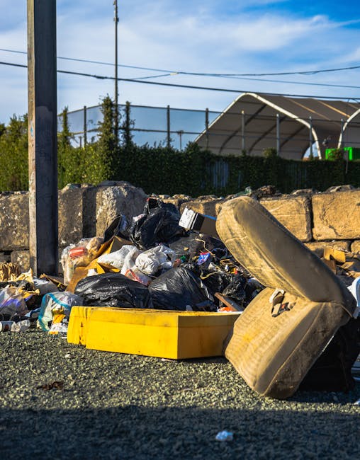 A collection of discarded waste materials and rubbish on a gravel surface, including several black plastic trash bags filled with waste, a yellow plastic container, a worn and stained car seat, and scattered debris. The waste is piled against a low stone wall, with some items leaning or partially resting on it. In the background, there is a fence with green shrubbery, utility poles with electrical wires, and a large industrial or storage tent with a curved roof, all under a partly cloudy sky with blue tones. The scene depicts an outdoor area where private waste collection or rubbish removal services, such as those offered by Rubbish Removal Uxbridge, might be required to clear the accumulated waste before public or environmental concerns arise.