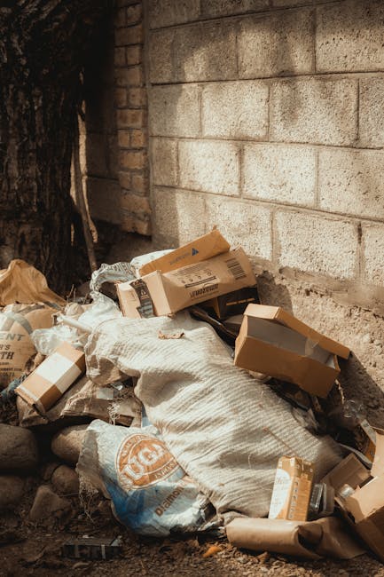 A pile of miscellaneous waste materials is gathered in front of a light-colored brick wall at the base of a large tree trunk. The waste includes cardboard boxes, some flattened and others partially intact, with visible printed labels and brown kraft finishes. There are also large plastic bags, some with printed logos, filled with unidentifiable rubbish, and one bag appears to be made of woven material. The boxes and bags are roughly stacked, creating a disorganized collection of refuse near the ground. The scene is outdoors, with a mix of natural and constructed elements; the brick wall behind the rubbish indicates an urban or residential environment, while the nearby tree adds a natural element. The lighting suggests daytime, with soft shadows cast on the wall and ground. This image reflects the type of unmanaged waste that can be addressed through private rubbish removal services like those offered by Rubbish Removal Uxbridge, supporting sustainable and responsible waste disposal practices outside of standard council collections.