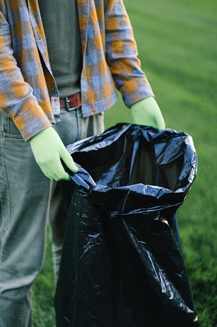 A person standing outdoors on a grassy area, holding open a large black rubbish bag lined with a metallic inner layer. The individual is wearing a plaid shirt in shades of yellow, grey, and blue, along with green disposable gloves, with the shirt partially unbuttoned to reveal a green T-shirt underneath. The person's jeans are visible, secured with a brown belt. The rubbish bag appears to be empty or contains minimal waste, and the scene suggests a waste collection or disposal activity, typical of private rubbish removal services. The background features out-of-focus greenery, indicating a park, garden, or similar outdoor setting. The lighting is natural, providing clear visibility of textures such as the crinkled plastic bag, the plaid fabric, and the gloves. The image aligns with themes of waste management, on-site rubbish clearance, and environmentally responsible disposal practices, highlighting methods often used in independent rubbish collection in residential or outdoor environments.
