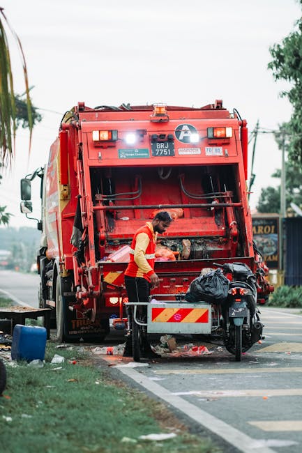 A worker wearing a yellow safety vest and dark trousers is operating a red rubbish collection vehicle, which is positioned on the side of a paved street. The vehicle's rear hatch is open, revealing mechanical components and a partially visible loading area. The worker appears to be managing waste, with a black plastic bag placed on a small metal cart attached to the vehicle. Behind the worker, a motorbike is secured with a rear cargo rack, and some scattered debris, including small pieces of trash, is visible on the ground beneath the vehicle. In the background, there are trees, utility poles with wires, and a building with signage. The scene is lit by natural daylight, with a partly cloudy sky overhead, and the setting looks like an urban or suburban street that may require private waste collection services, such as those provided by Rubbish Removal Uxbridge, to manage local rubbish effectively and efficiently.