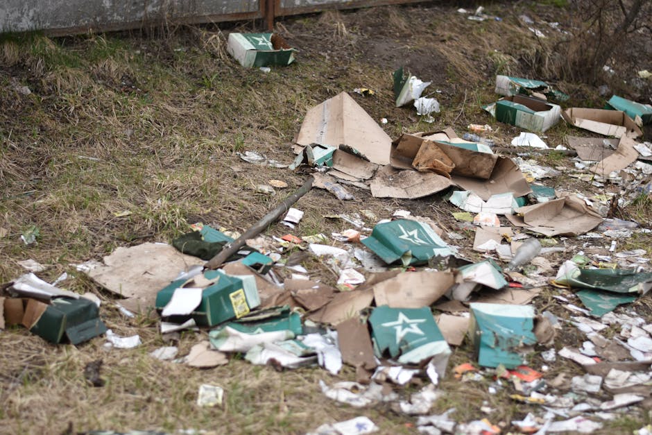 A section of an outdoor area adjacent to a grassy verge with scattered debris including flattened and torn cardboard boxes, some with printed logos and labels, mixed with torn paper and plastic packaging. The cardboard appears weathered with some flattened and partially crumpled, exhibiting brown, beige, and green tones with visible textures such as ridges and corrugation. There is a metal rod or pole lying among the rubbish, partially obscured by the debris. The ground underneath consists of uneven soil and sparse grass, with small patches of greenery visible near the edges of the scattered waste. In the background, a brick wall or fence boundary can be seen, and the overall environment suggests neglected or improperly disposed rubbish, illustrating the type of discarded waste that private rubbish removal services like Rubbish Removal Uxbridge might assist with in managing the cleanup of such fly-tipped or unmanaged waste.